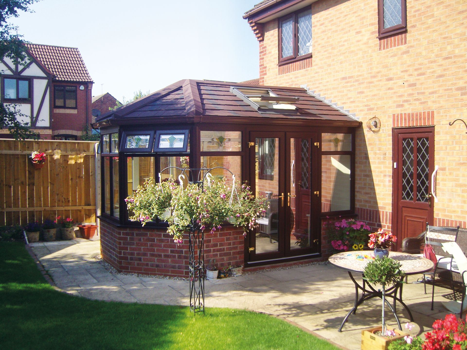 Brown conservatory attached to a brick house, with a patio, flowers, and lawn.