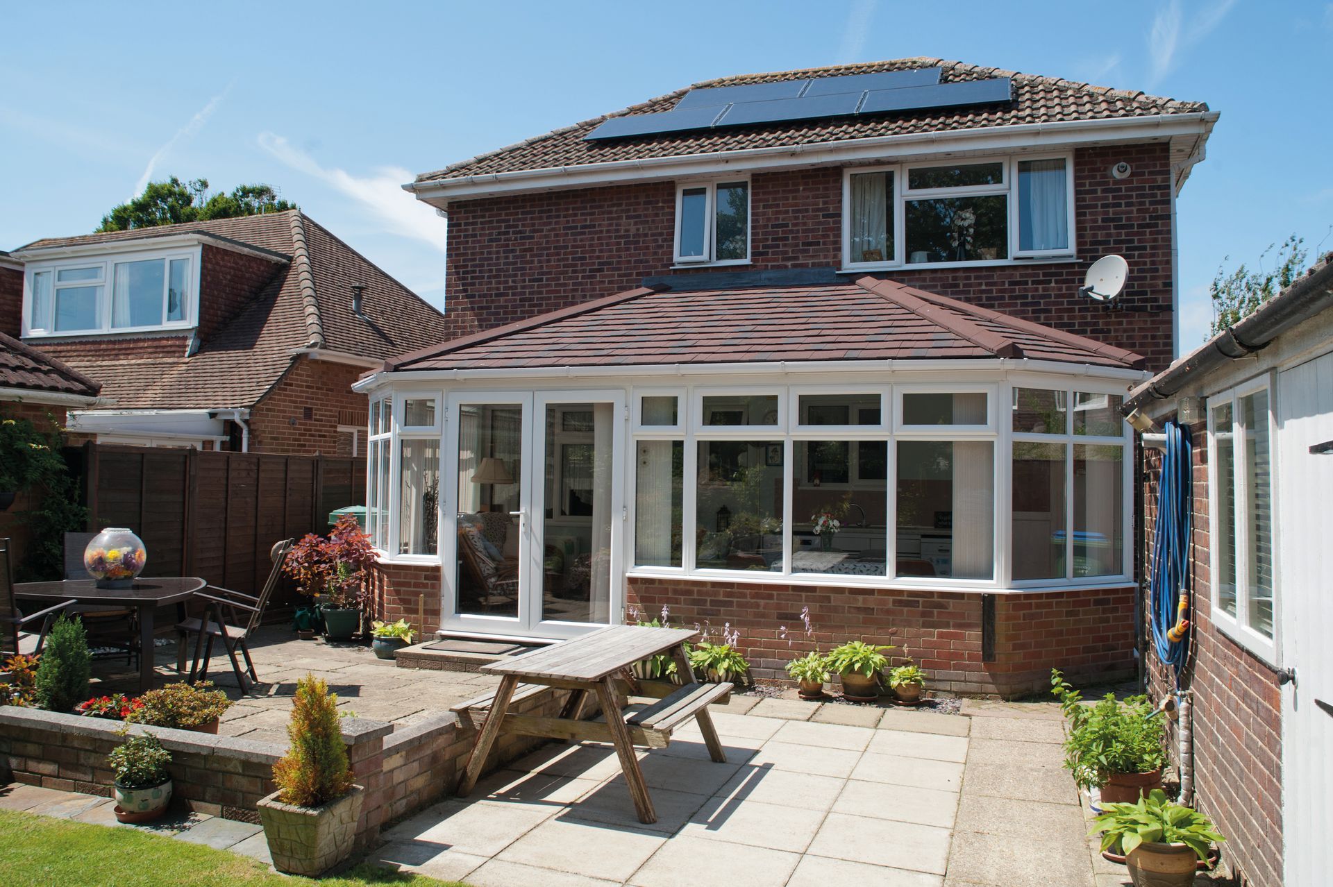 Backyard patio with brick home, conservatory, table, plants, sunny day.