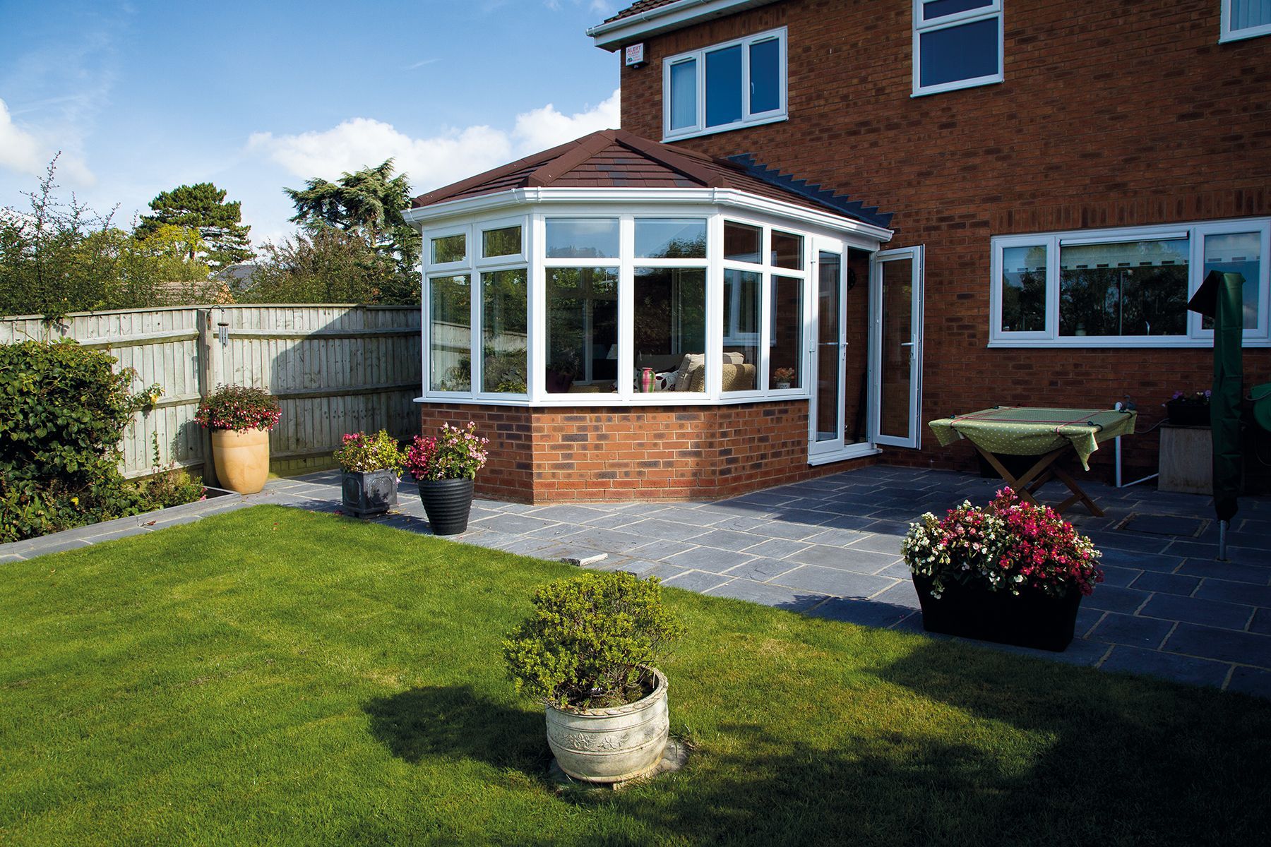 Brick house with a conservatory on a patio, lush green lawn, and potted flowers.