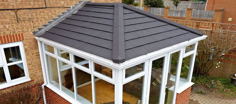 A conservatory with a dark gray roof and white window frames, built on a brick base.