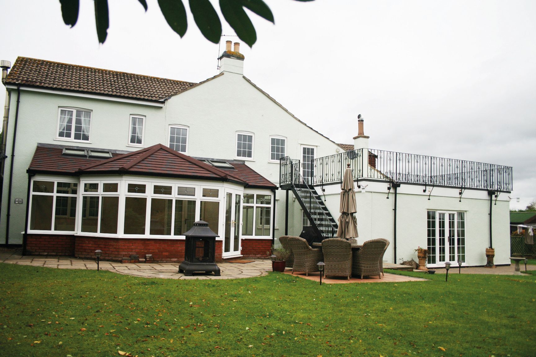 Large white house with a brick-roofed conservatory, patio, and balcony on an overcast day.