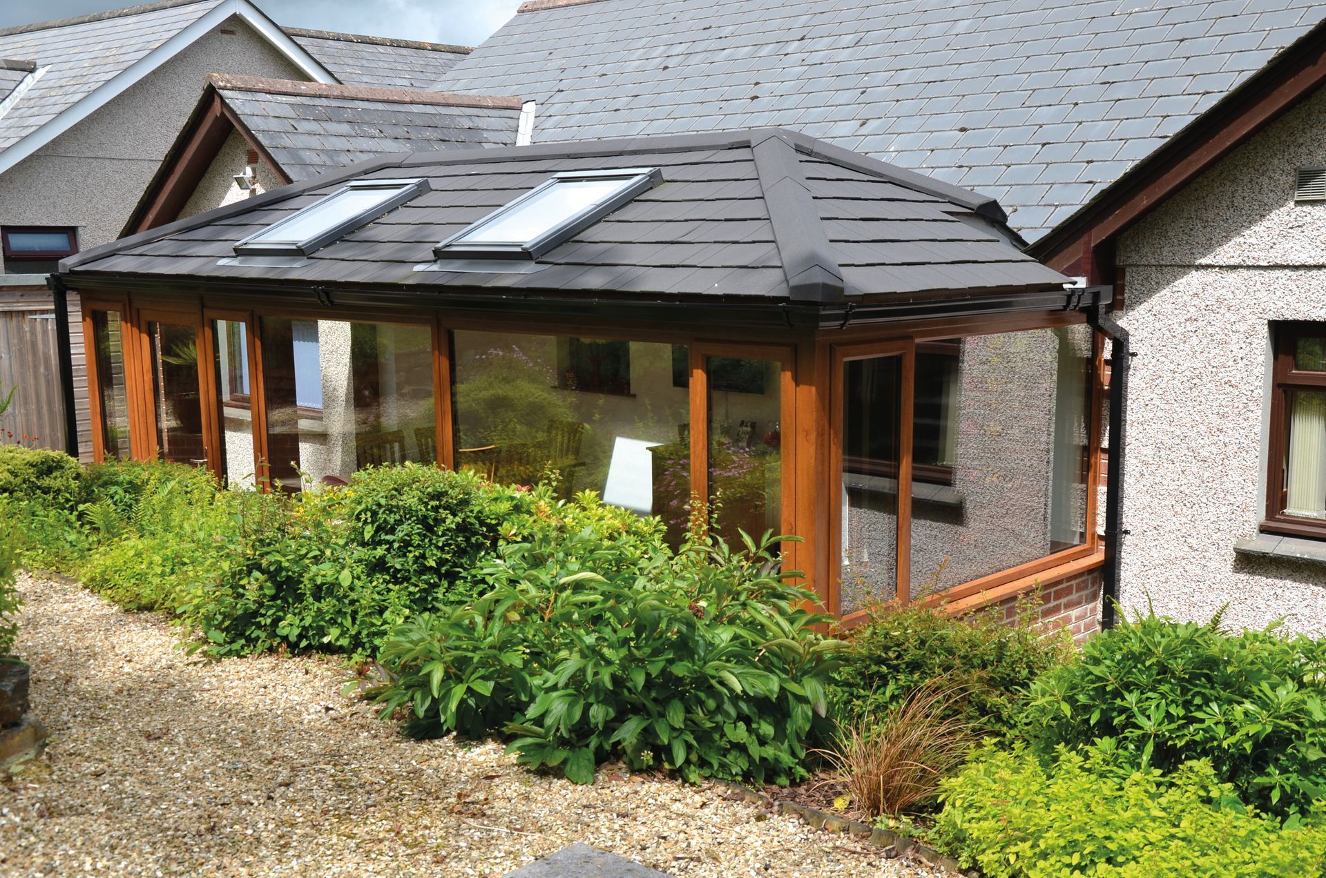 Sunroom with glass walls, wooden trim, and dark tile roof, flanked by bushes, set alongside a house with gray siding.
