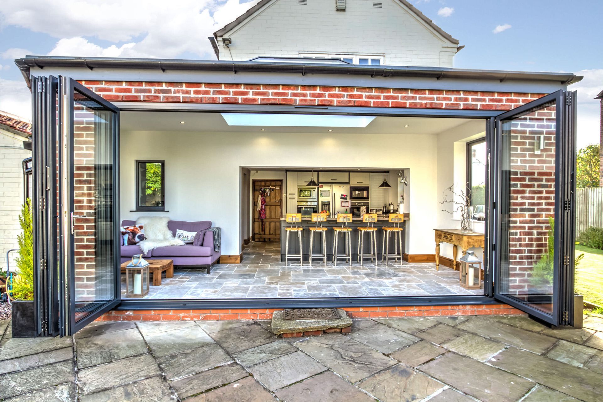 Rear view of a brick-framed extension with bi-fold doors open to a patio. Interior includes a kitchen and seating area.