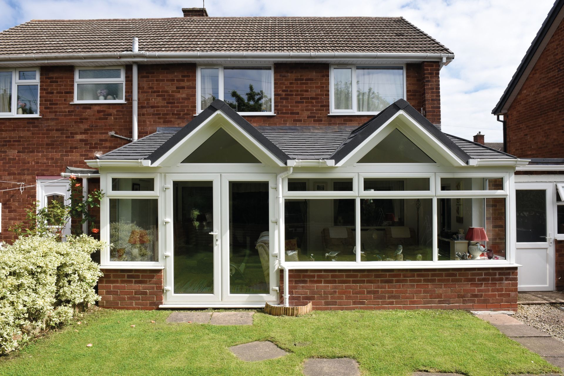A brick and white-framed conservatory with a grass lawn, two-story red brick house behind.