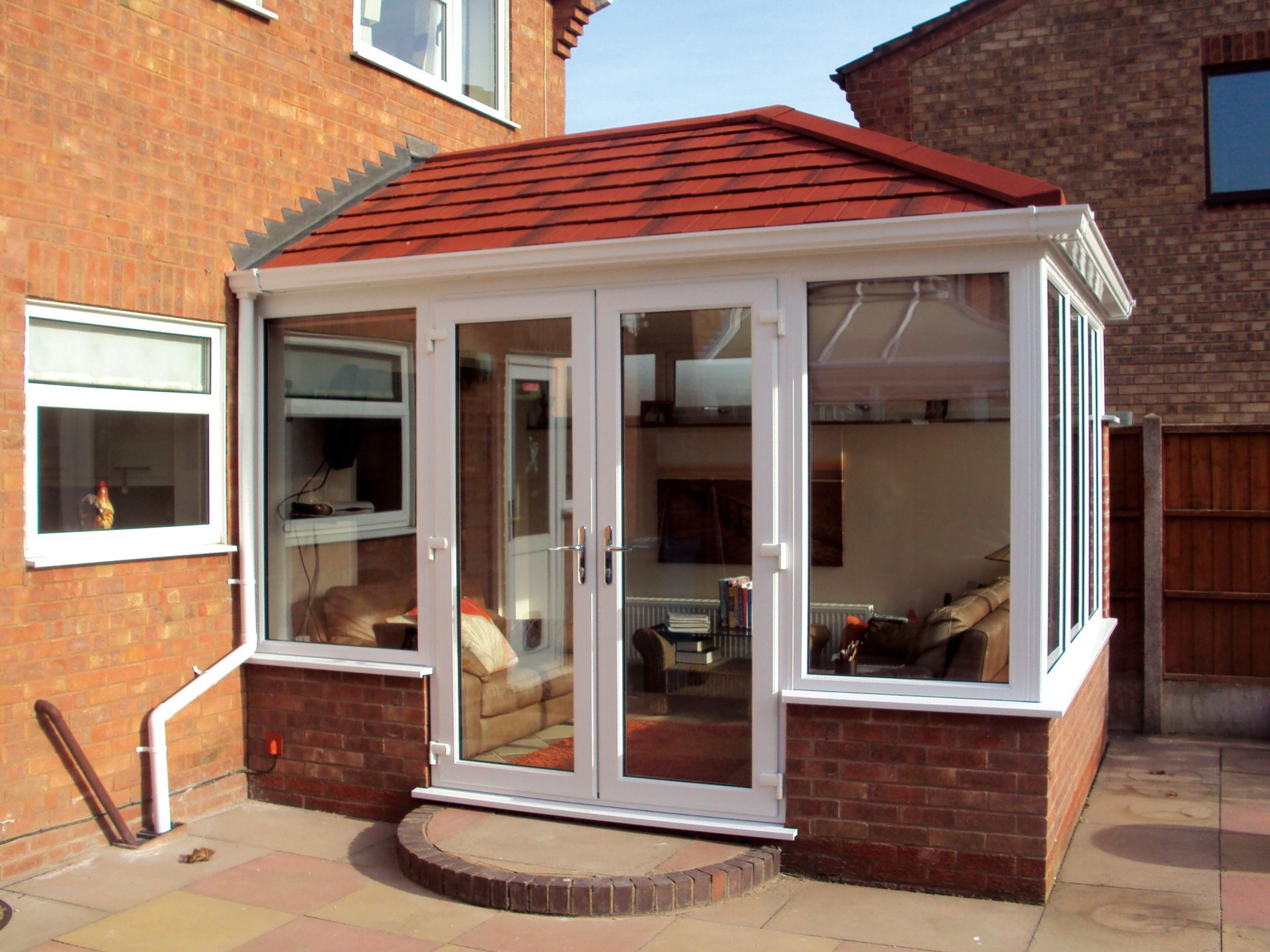 A sunroom with white frames, glass windows, red roof, and brick base attached to a brick house.