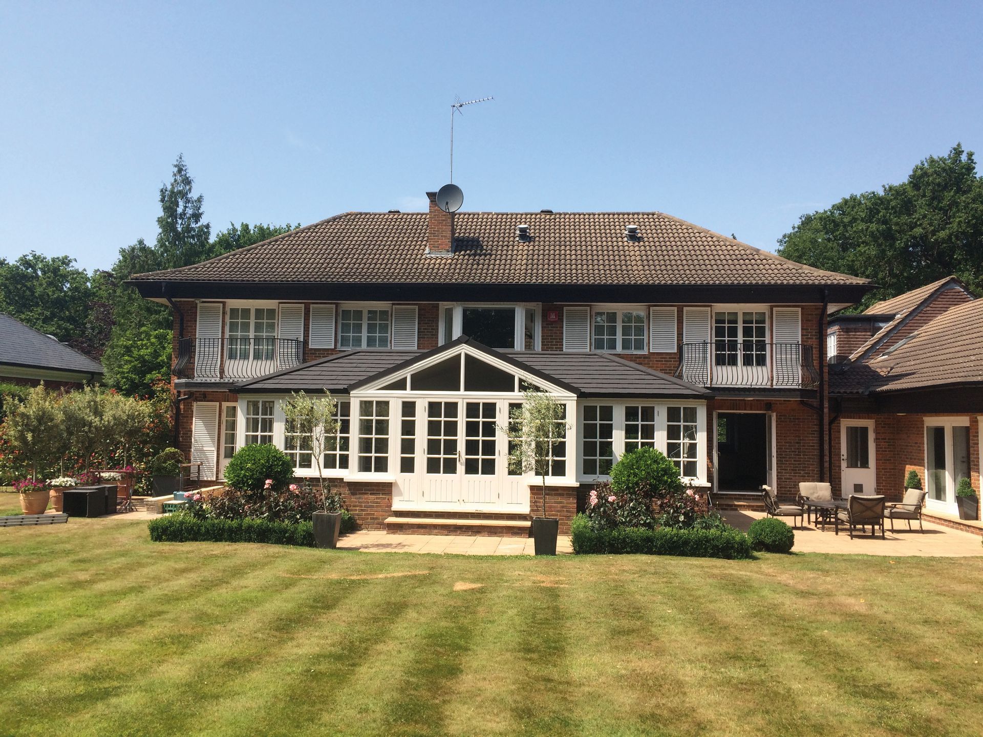 Two-story house with white shutters, brick exterior, tiled roof, glass-walled porch, and manicured lawn on a sunny day.