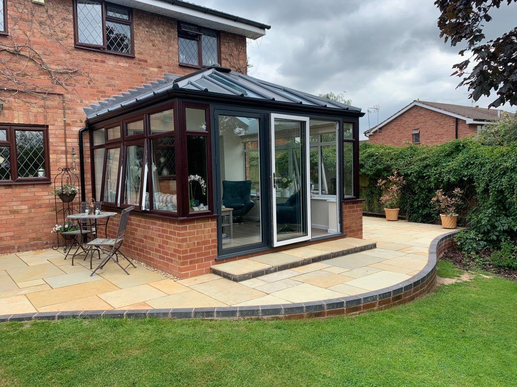 A conservatory with a brick base, glass walls, and dark roof, adjoining a brick house with a stone patio.