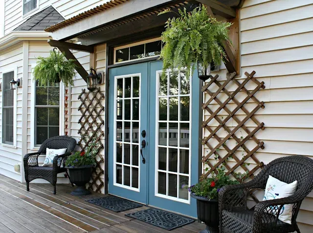 Blue double doors on a house with fern-filled pots, wicker chairs, and a wooden trellis.