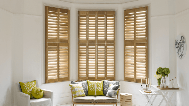 A bright living room with bay windows featuring wooden shutters; a small sofa with pillows sits below.