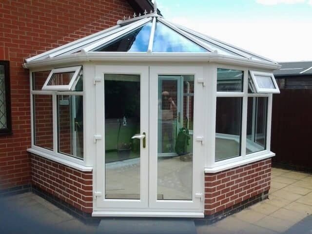 White-framed conservatory with glass windows and doors, set on a brick base, against a red brick wall.