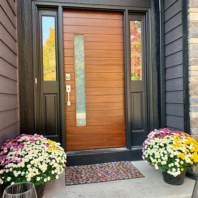 Modern brown front door with sidelights, welcome mat, and flower pots on a concrete porch.