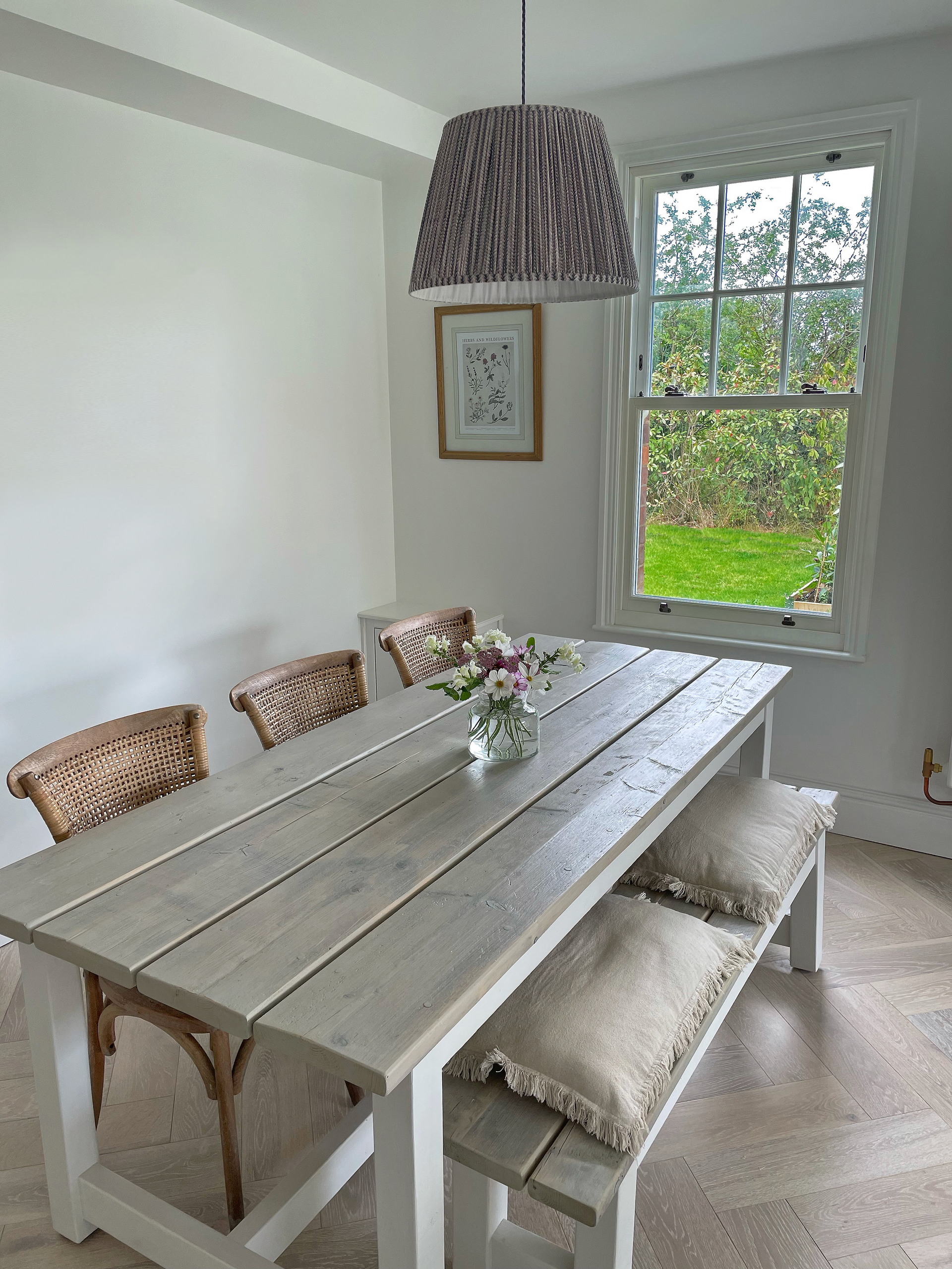 Dining room with light gray wood table, benches, woven chairs, and a window overlooking a green yard.