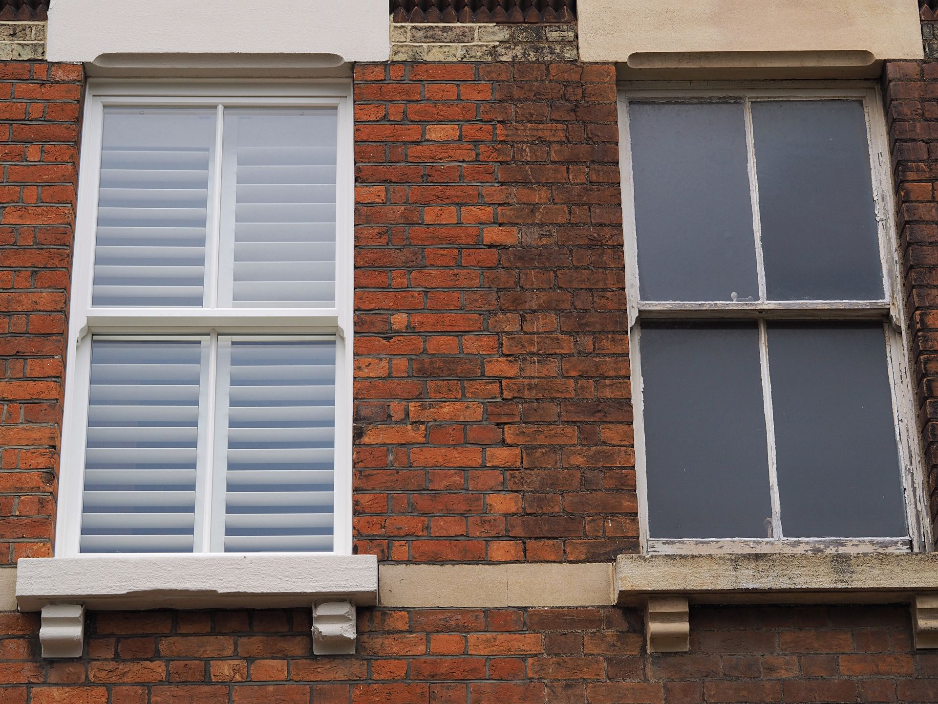 Two windows in a brick building. One with white shutters, the other dark with white trim.
