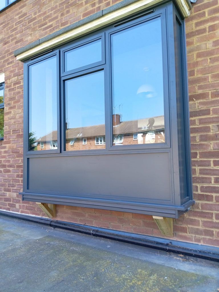 Bay window with gray frame, reflecting sky and buildings, installed on a brick wall, supported by wooden brackets.