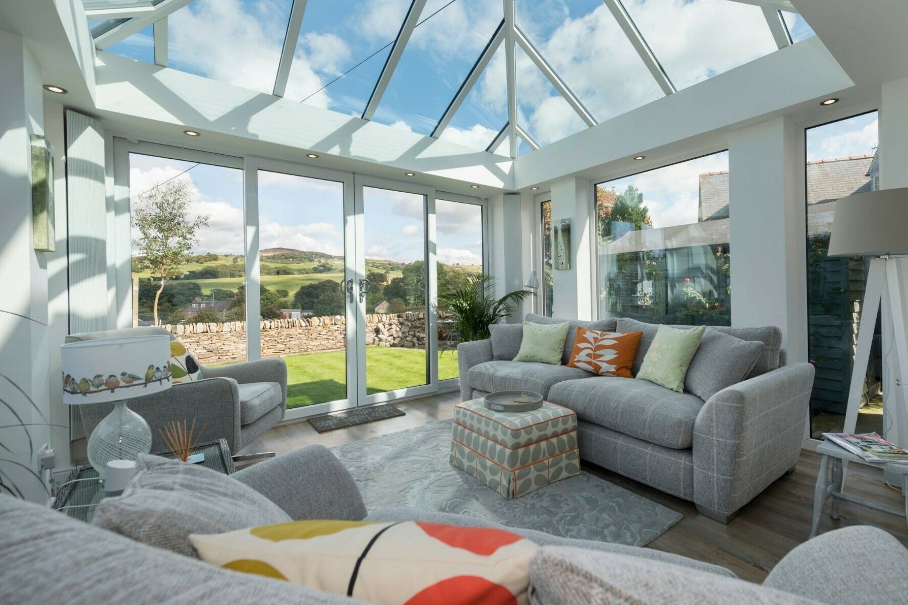 Sunroom with glass roof, gray couches, and a view of a green yard and blue sky.