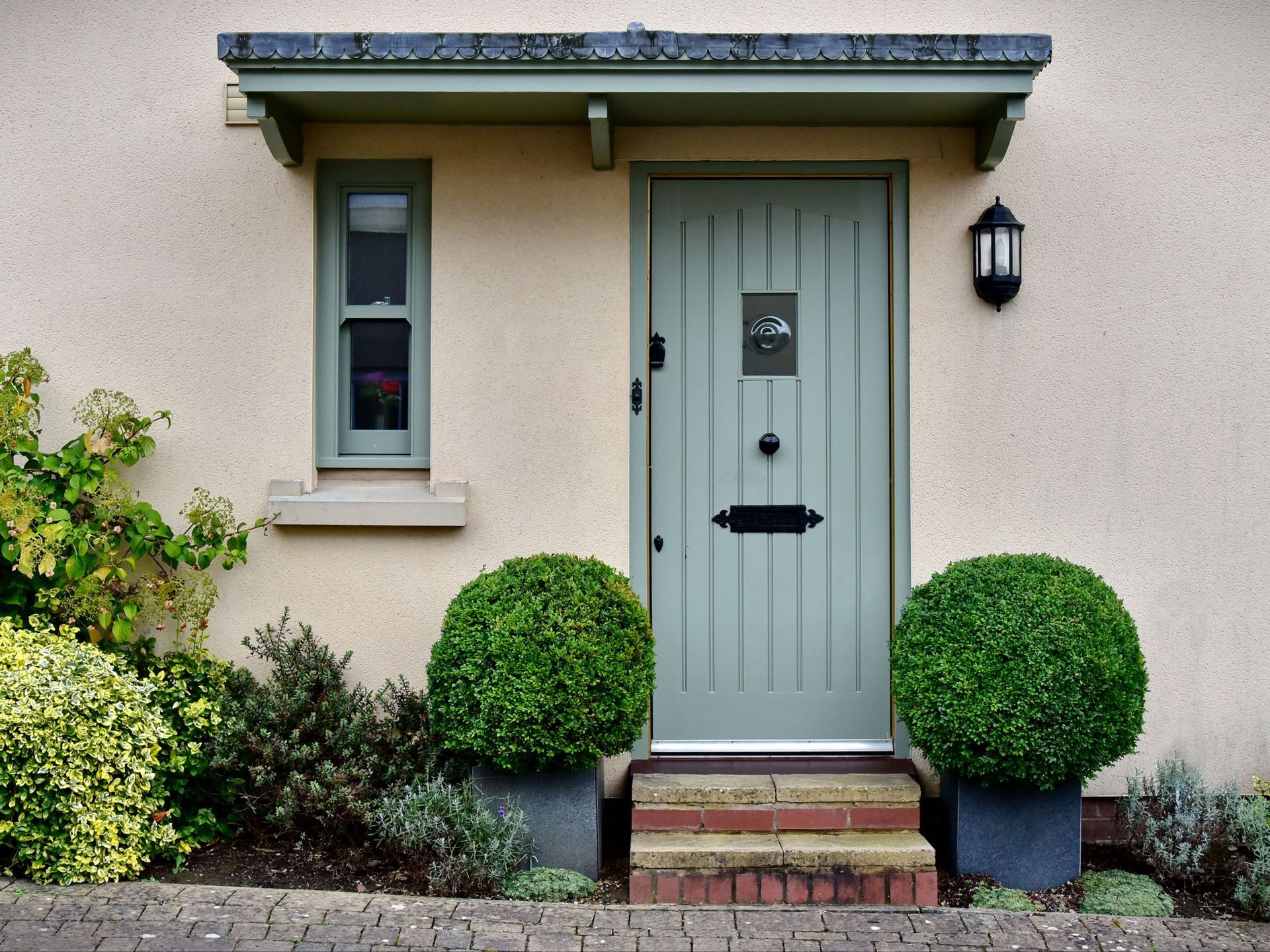 A light green door with a small window and round bushes in front.