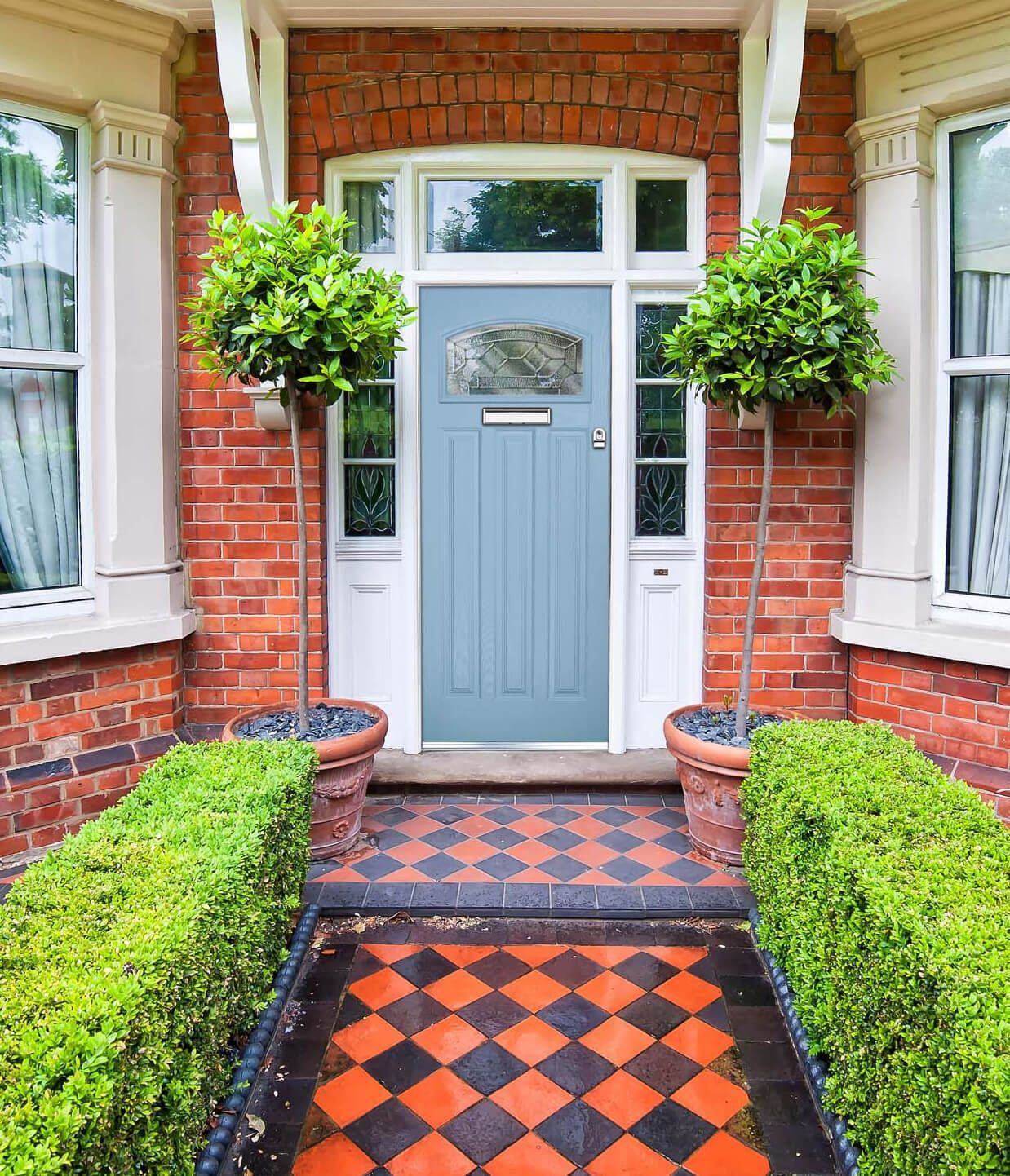 Blue door with two topiary trees and checkered brick path leading to a red brick house.