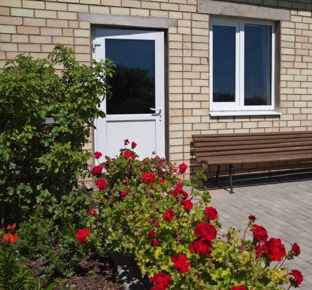 Brick building with white door and window, bench, and red geraniums in a garden.