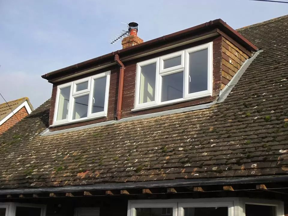 Dormer with two white-framed windows on a weathered, tile roof. Brick siding, antenna, and a clear sky.