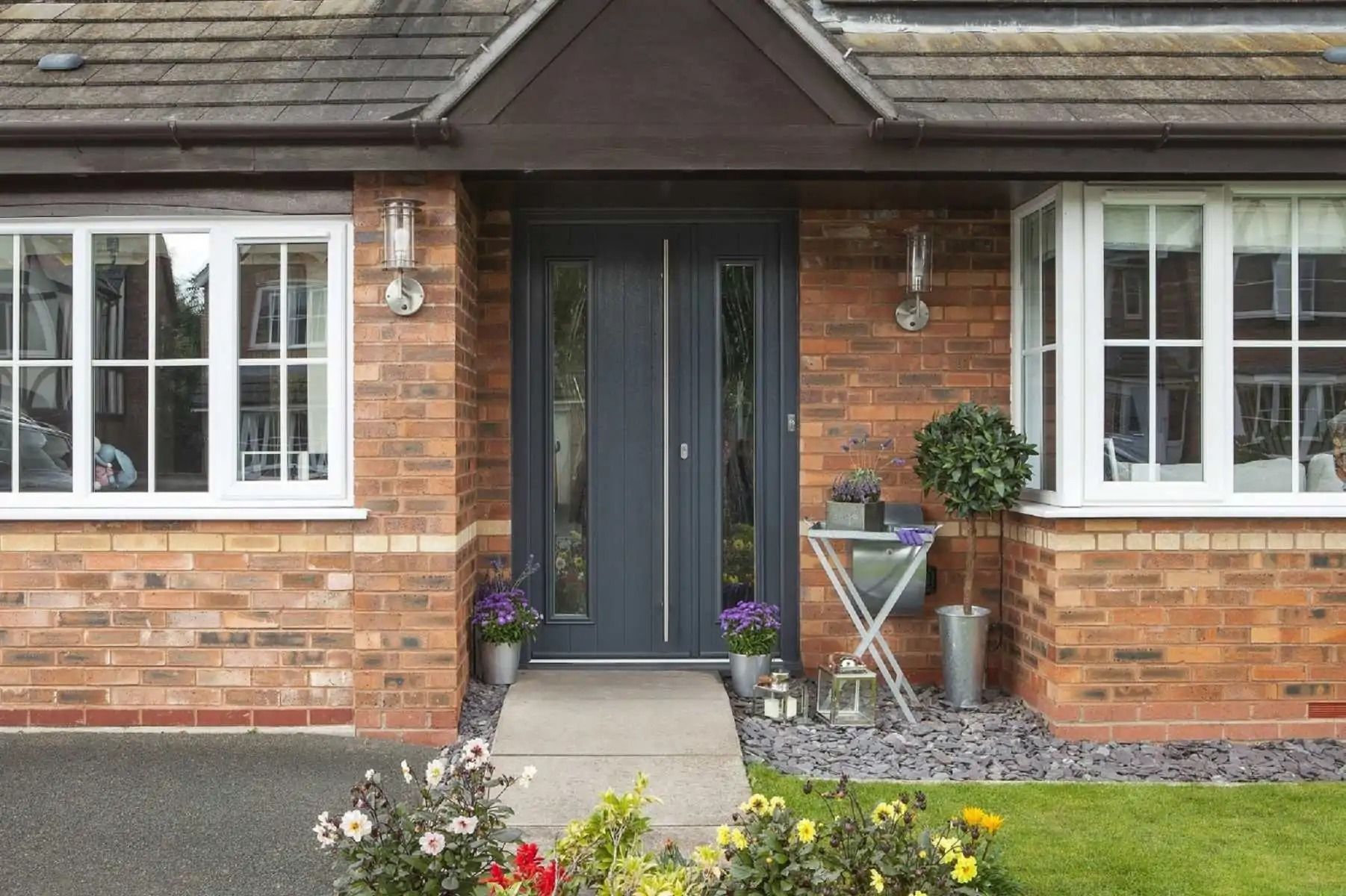 Gray front door with sidelights on brick home with white windows; flowers and a small table on the porch.