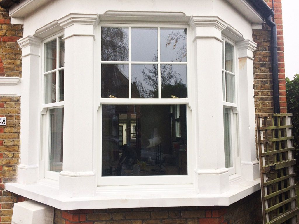 White bay window on a brick building. Large glass panes reflect outdoors; small garden visible on the right.
