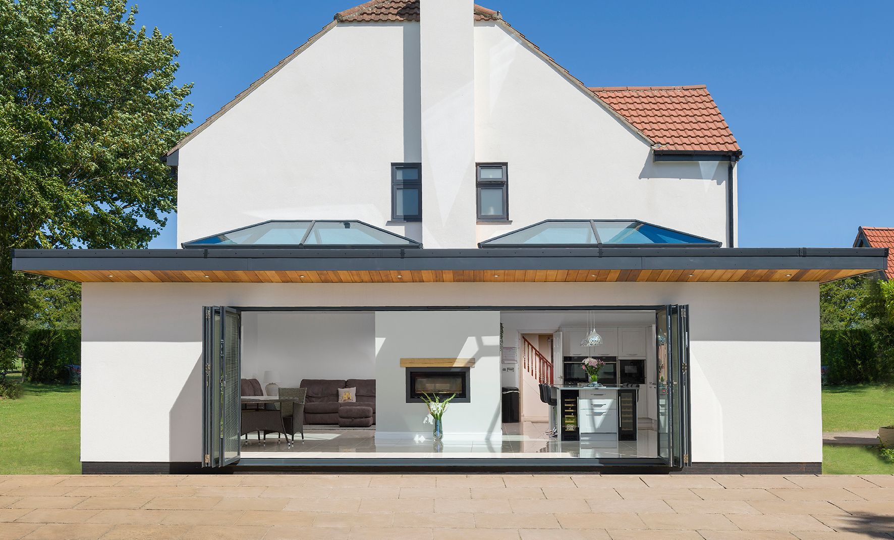 White house with a modern, glass-walled extension. Open folding doors reveal interior. Blue sky.