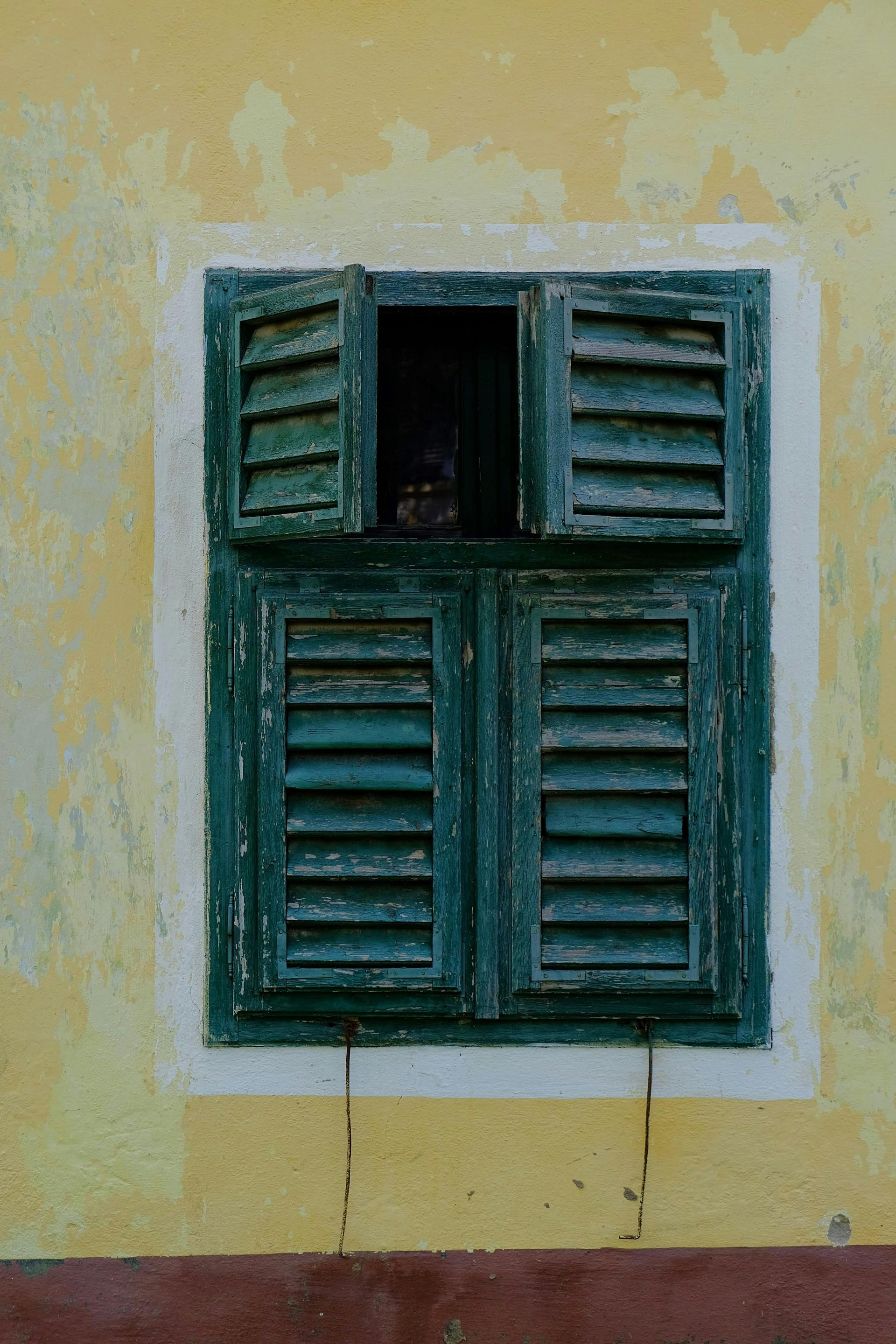 Green shuttered window on a yellow wall, one shutter open, revealing a dark interior.