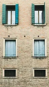 Four windows of varying sizes on a brick building. Top windows have teal shutters. Bottom have barred windows.