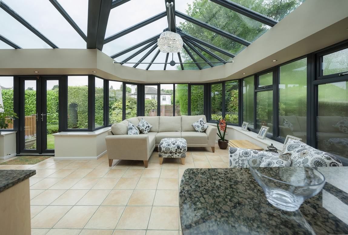 Sunroom with beige couch and tiled floor, surrounded by large windows overlooking a green garden.