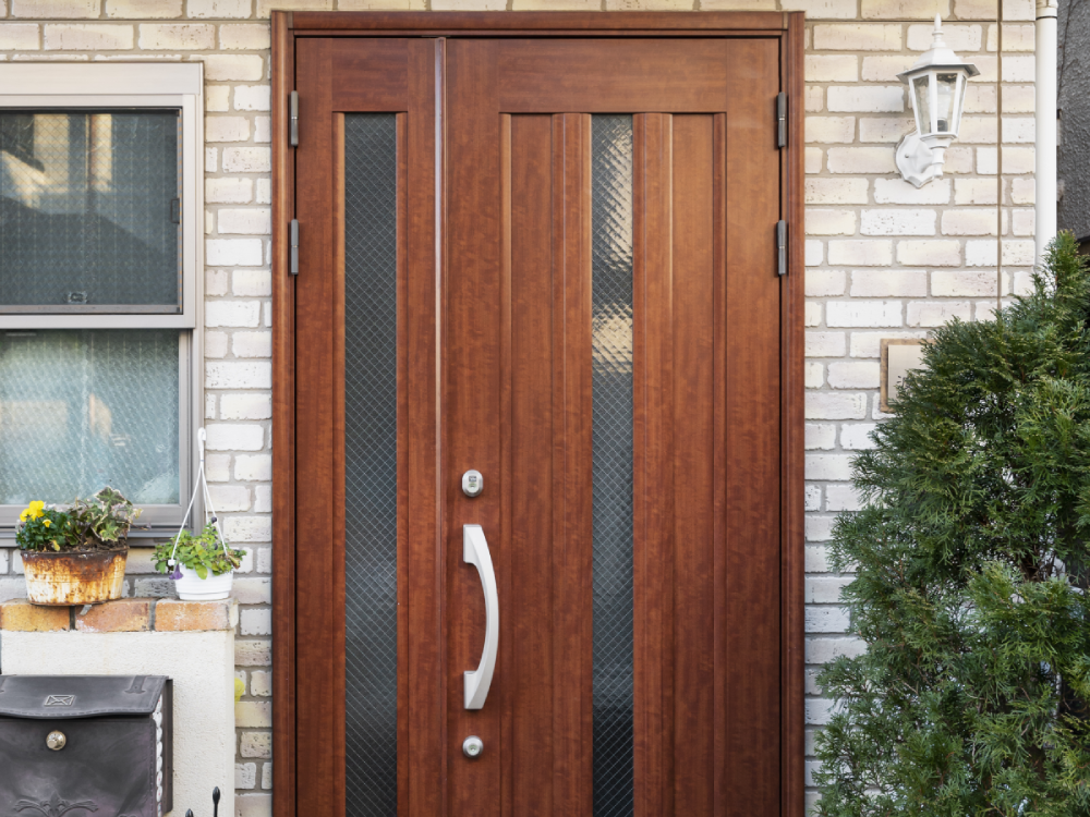 Double wooden front door with glass panels, set in brick wall with adjacent window and plant.