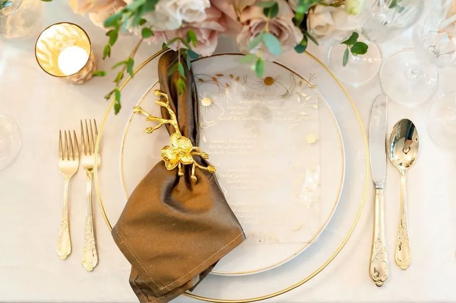 Elegant place setting with gold-rimmed plates, gold cutlery, a brown napkin, and floral decor on a white tablecloth