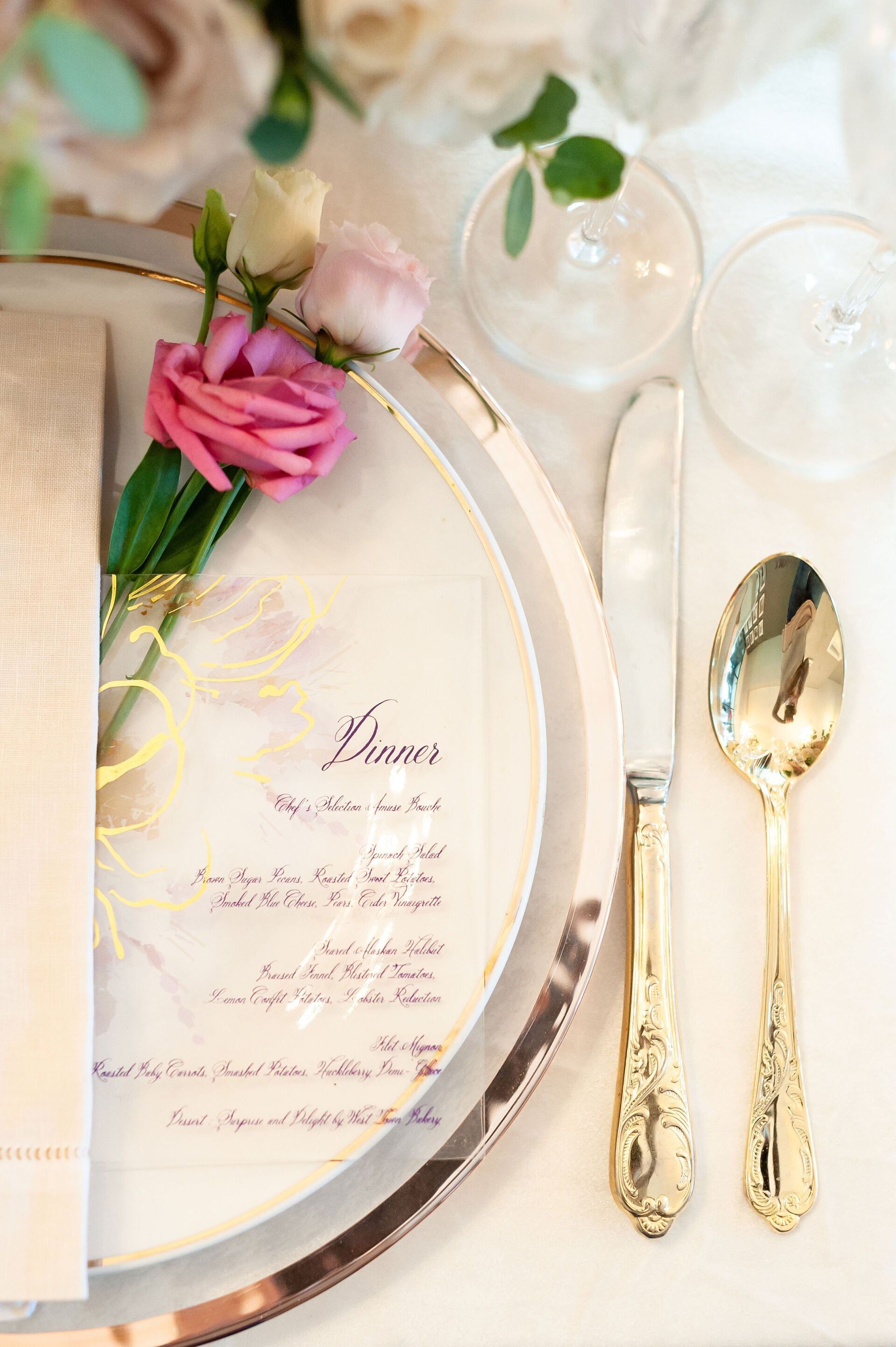 Elegant gold and white place setting with pink flower, menu card, and glassware on a table