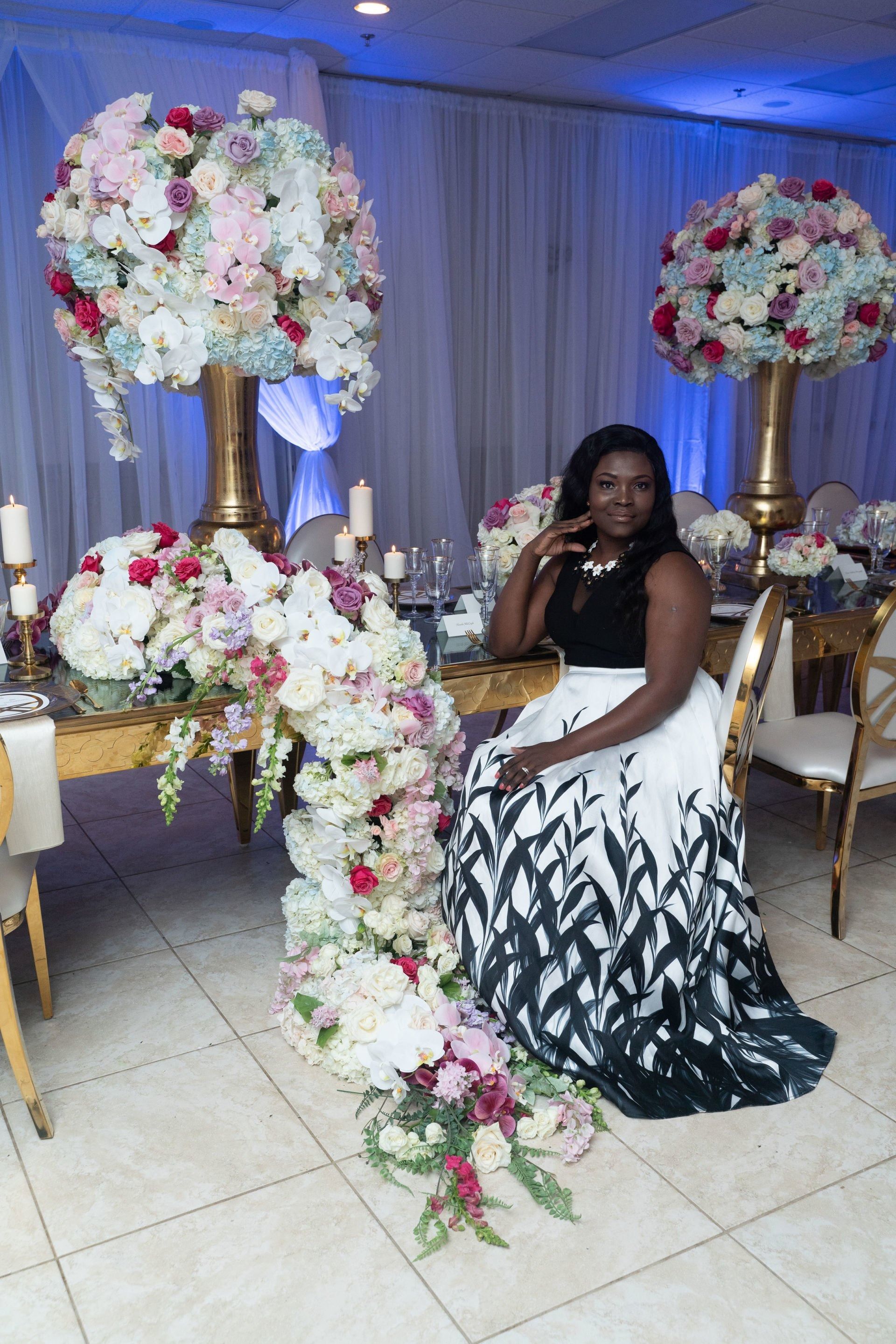 Woman seated at a decorated event table beside tall white-and-pink floral arrangements, wearing a black-and-white gown.