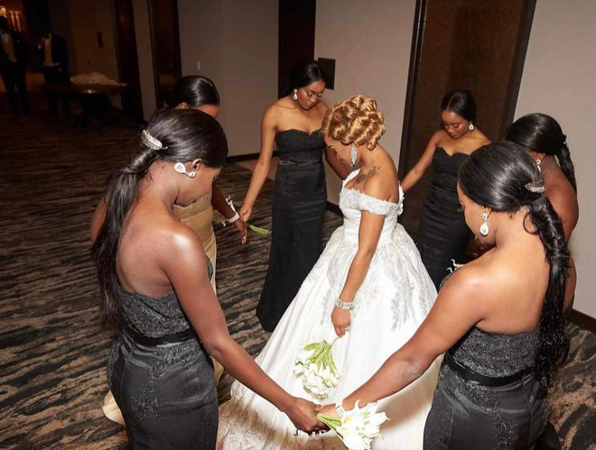 Bride in white gown stands with bridesmaids in black dresses, holding hands in a circle indoors.