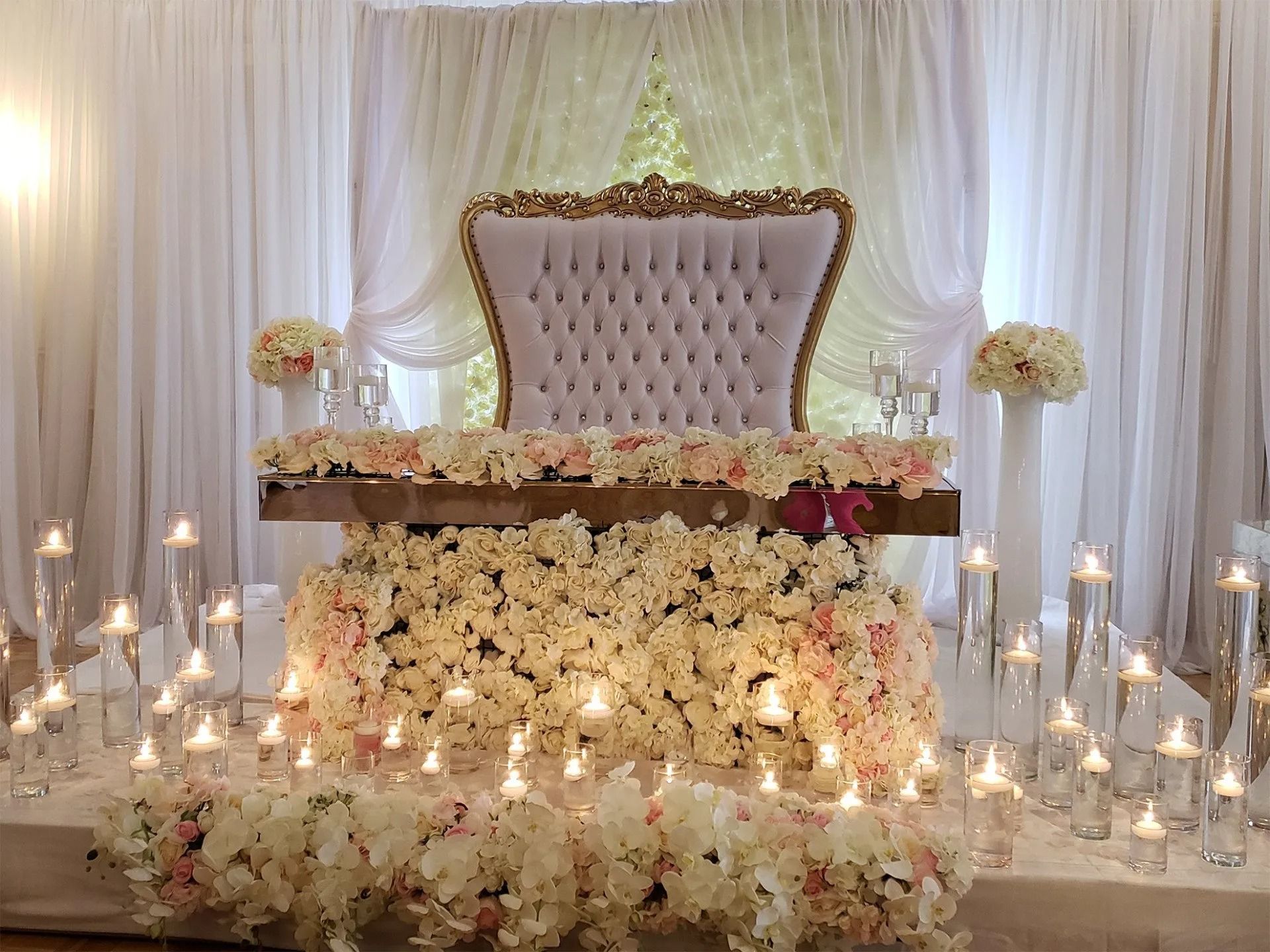 White floral wedding stage with tufted throne chair, candles, and draped curtains.