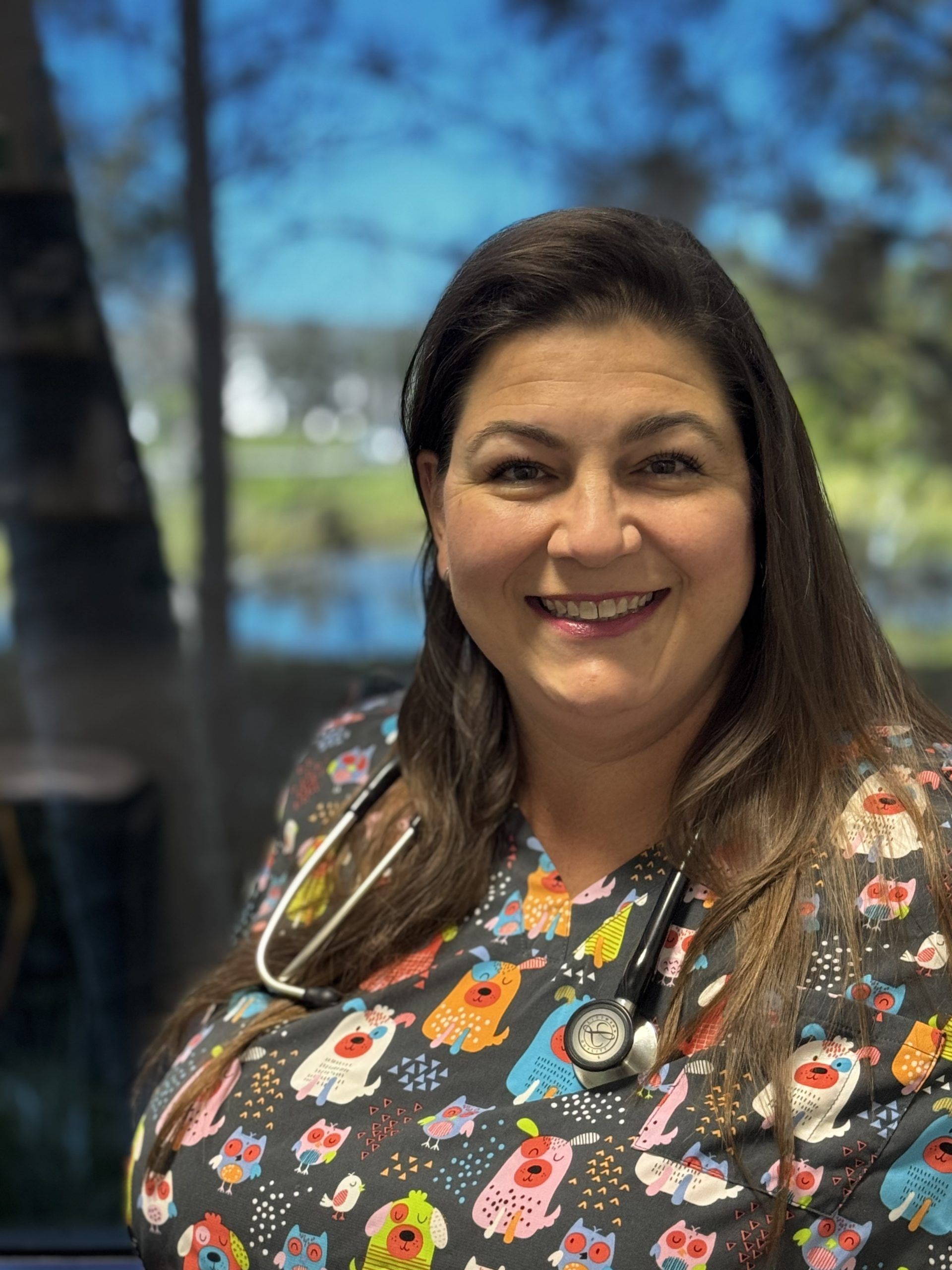 Smiling healthcare worker in patterned scrubs with a stethoscope, standing indoors by a window.