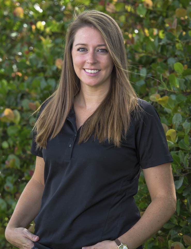 Smiling woman in a black polo shirt standing outdoors in front of green foliage.