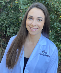 Smiling medical professional in a white coat standing outdoors in front of greenery