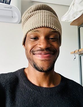 Smiling person wearing a beige knit beanie and black sweater indoors near a shelf.