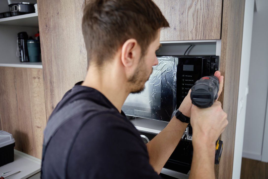 A person uses a power drill to install or repair a built-in kitchen appliance in a wood-paneled cabinet.