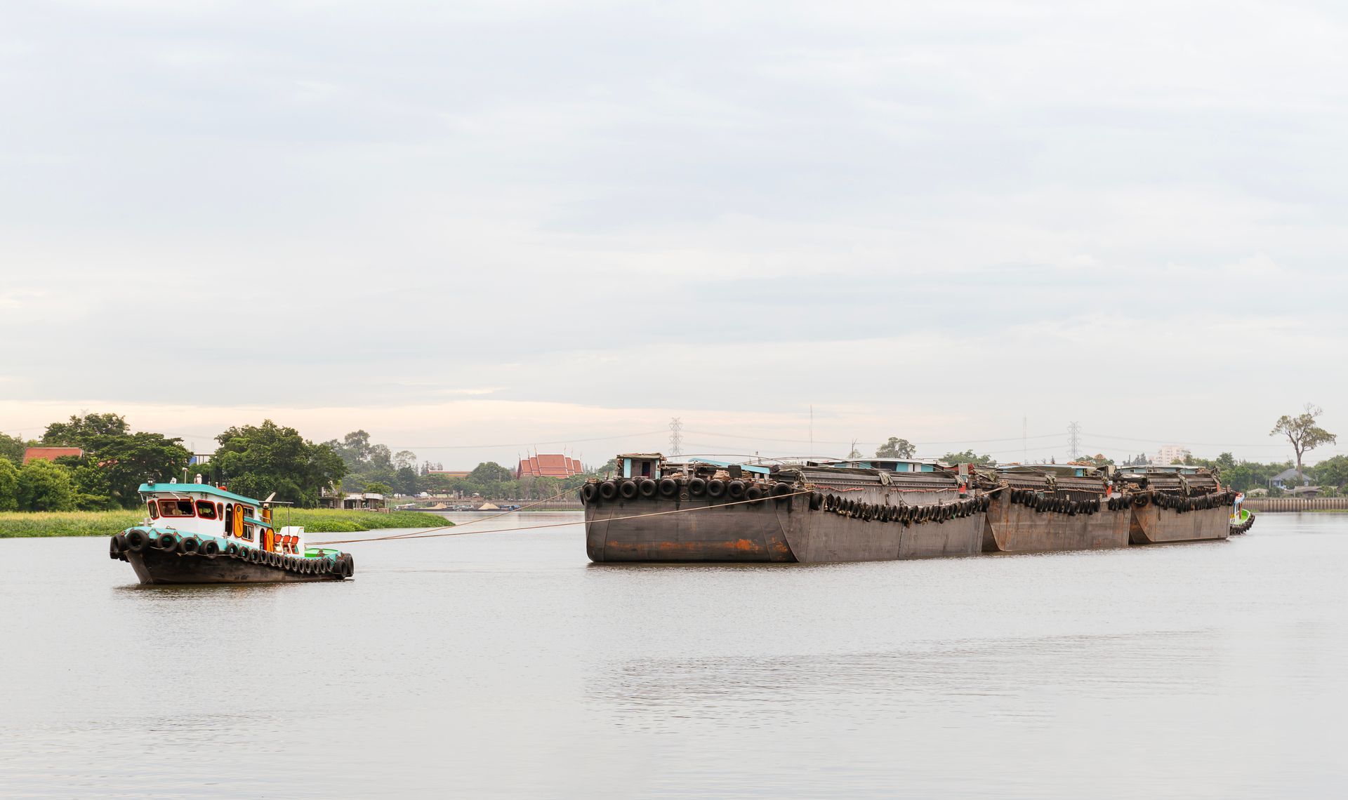 A tugboat pulls a long line of flat-bottomed cargo barges across a calm, wide river under a cloudy sky.