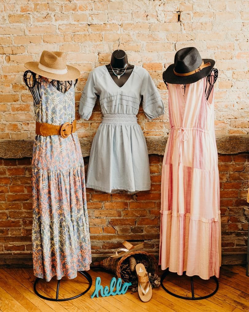 Three mannequins wearing dresses and hats are standing in front of a brick wall.