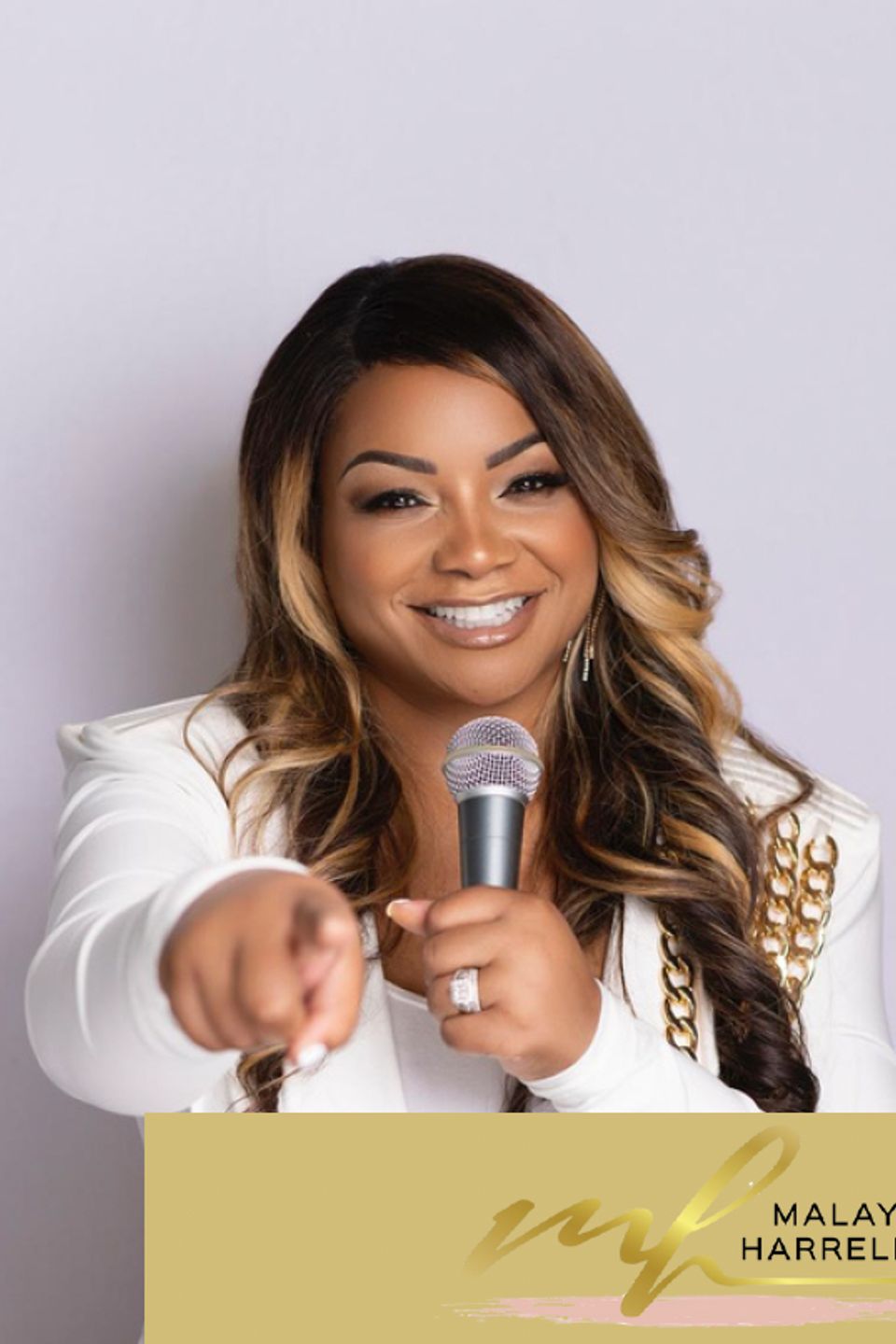 Smiling speaker holding a microphone and pointing toward the camera on a light background
