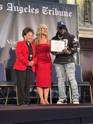 Three people onstage at an event, with one holding a certificate before a Los Angeles Tribune backdrop.