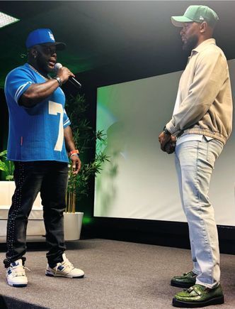 Two men on a stage facing each other, one holding a microphone in a blue jersey and cap.