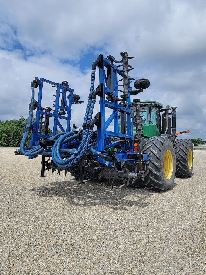 Blue agricultural sprayer attached to a tractor, parked on gravel under a cloudy sky.