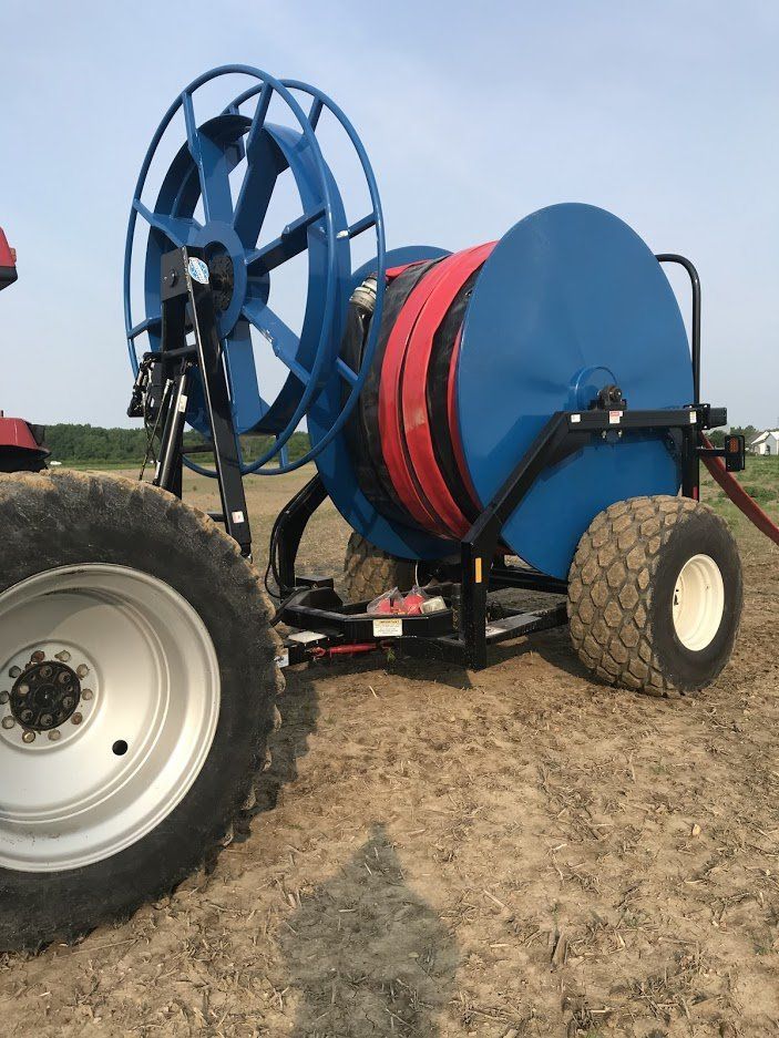 Blue farm irrigation reel trailer with large hose spool on a field, beside a tractor wheel