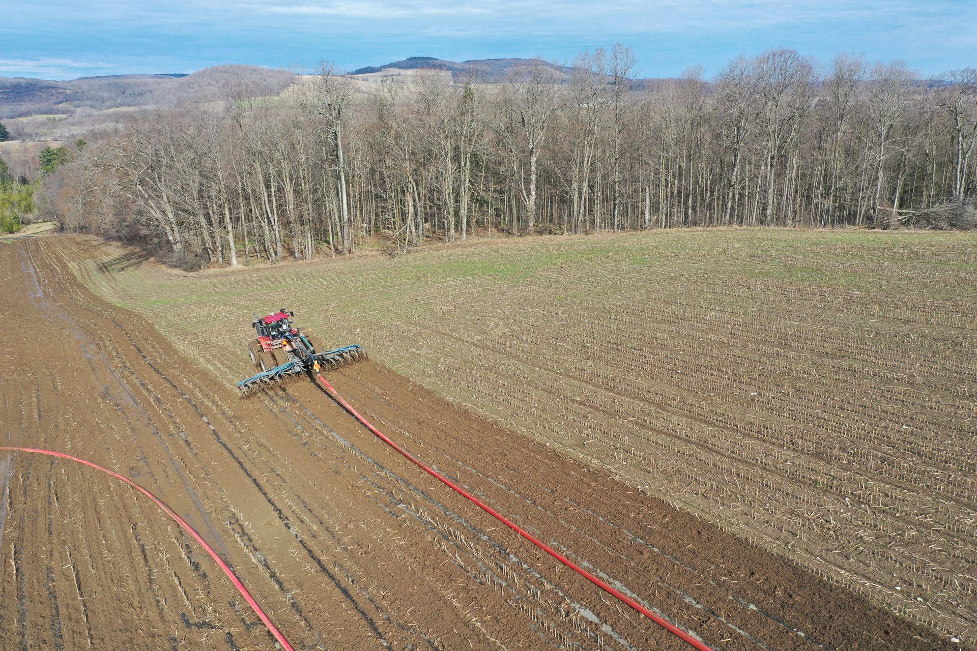 Aerial view of a red tractor plowing a brown field beside leafless trees and distant hills.