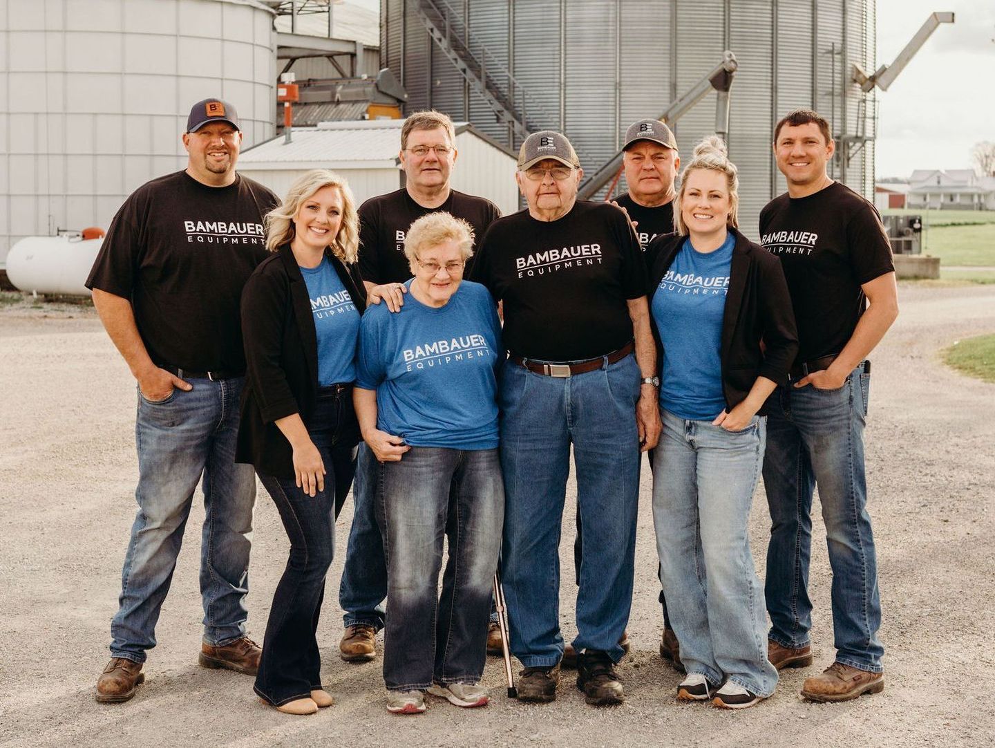 Seven people in black shirts and jeans stand smiling outdoors beside grain silos.
