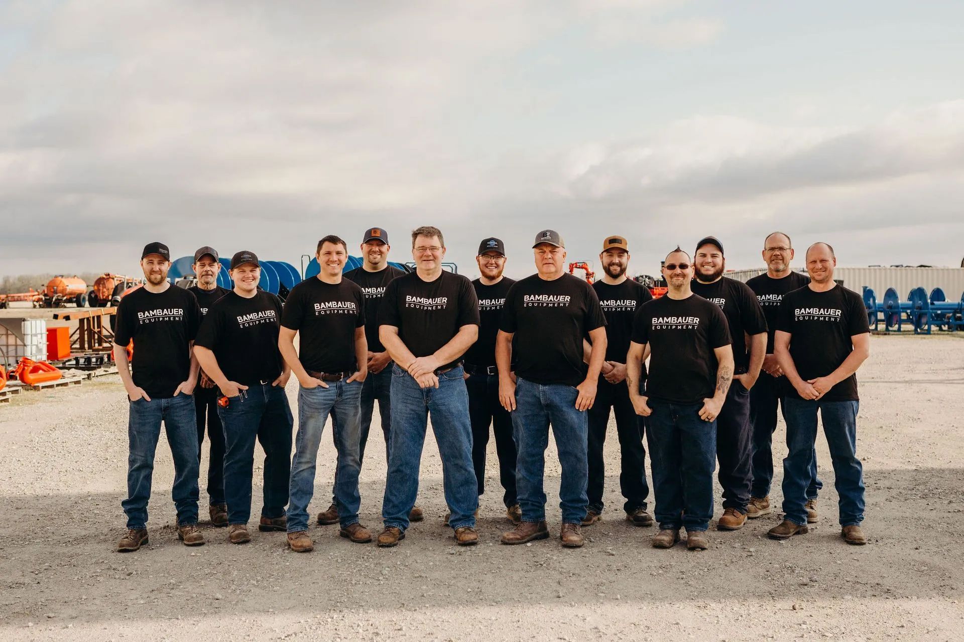 A group of people in black shirts and jeans standing outdoors on a gravel lot under a cloudy sky